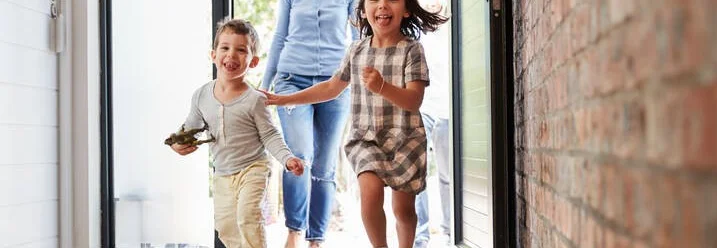 Zwei Kinder rennen lachend in ein Haus, gefolgt von einer Person.